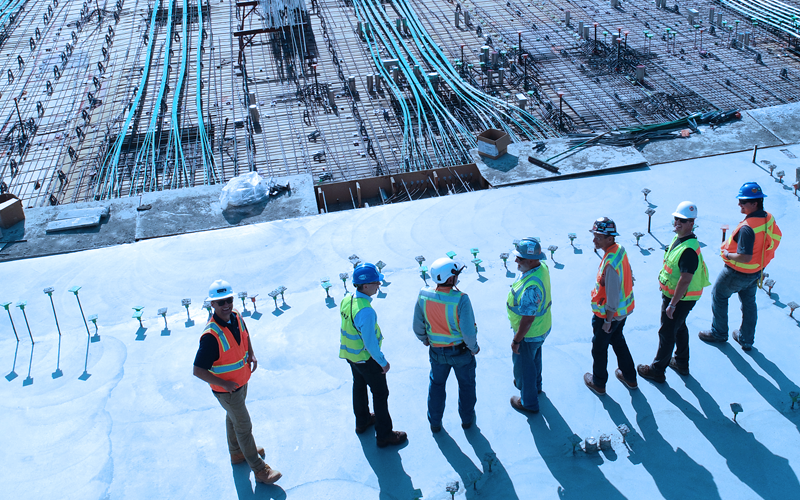 Group of construction workers standing on concrete formwork wearing yellow safety jackets. The sun is casting a long shadow behind them.